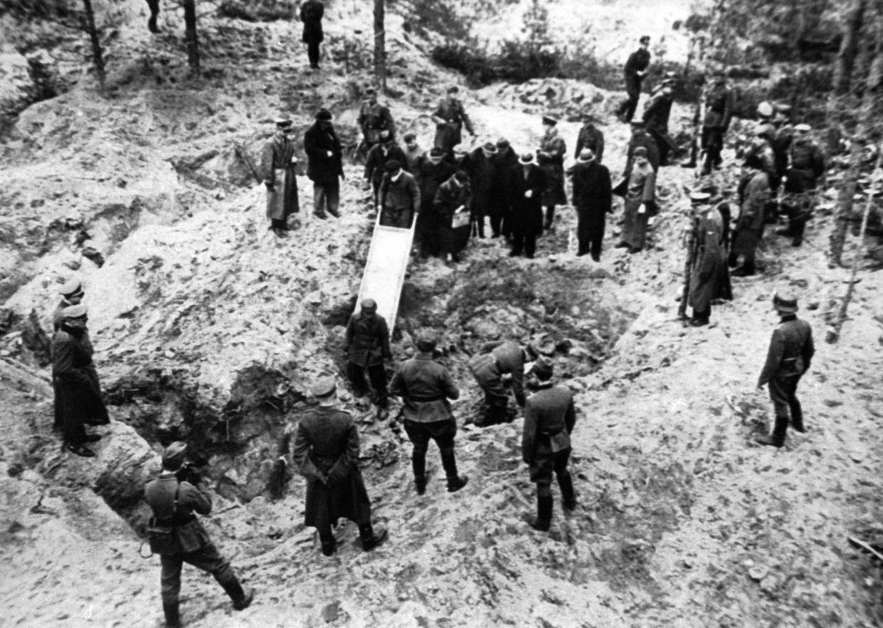 Black and white image, from above, of a mass grave in a clearing with dozens of people wearing long coats and hats or military uniforms standing by as two people hold a stretcher partway into a large hole in the ground.