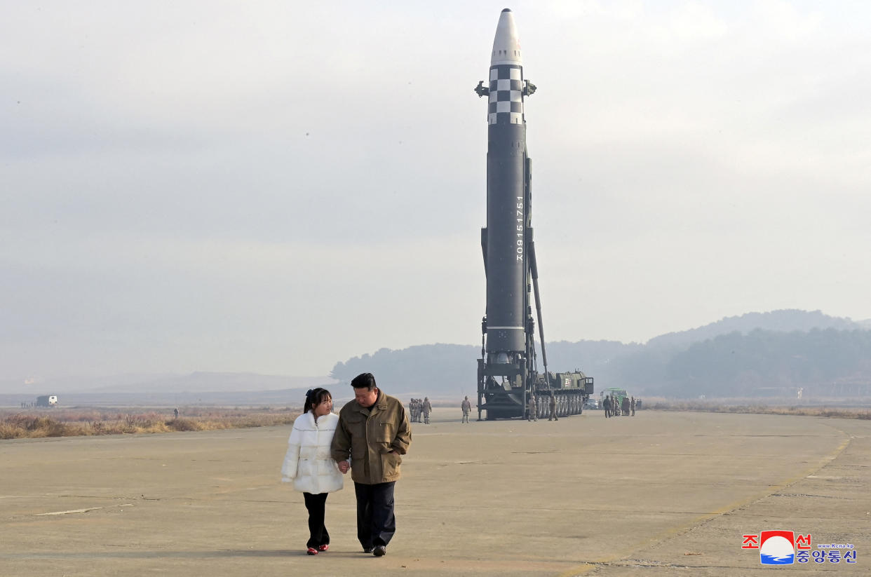 Kim Jong Un walks hand in hand across a flat expanse with a young woman in a white puffer jacket, a rocket with gray-and-white checkered midsection ready to launch behind them.