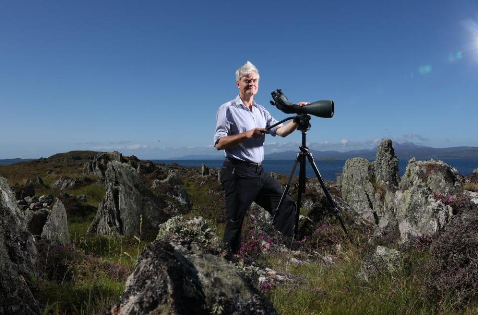 An older man stands on a rocky outcrop with a telescope.