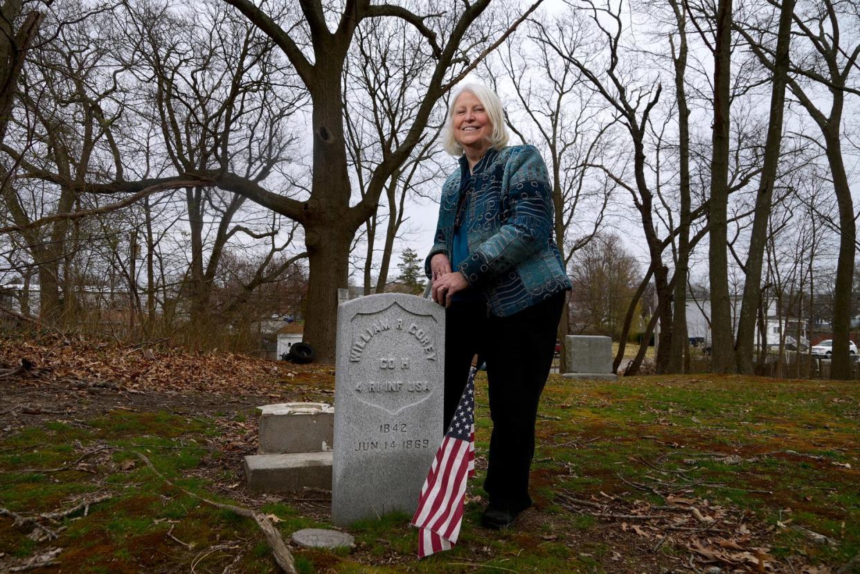 Pegee Malcolm, chair of the Rhode Island Historical Cemetery Commission, stands beside a new marker for Civil War veteran William Corey in a Warwick historical cemetery. On the ground behind the new marker is Corey's original stone, shattered by vandals.