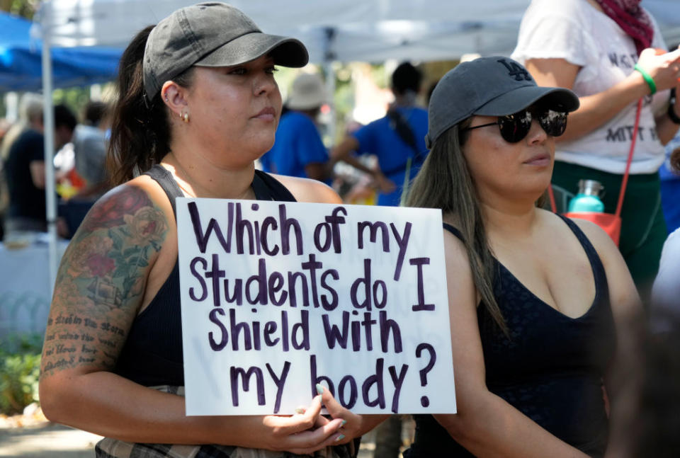 Two women at a protest hold a sign reading, "Which of my students do I shield with my body?" Both wear casual clothing and hats