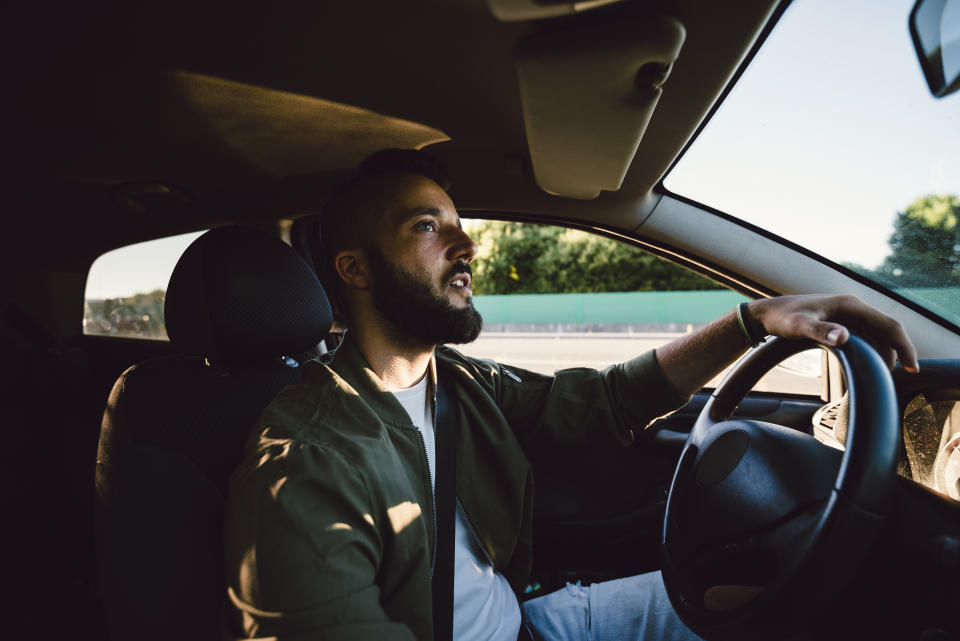 A man with a beard is driving a car, looking ahead with a thoughtful expression. He is wearing a casual jacket and a seatbelt