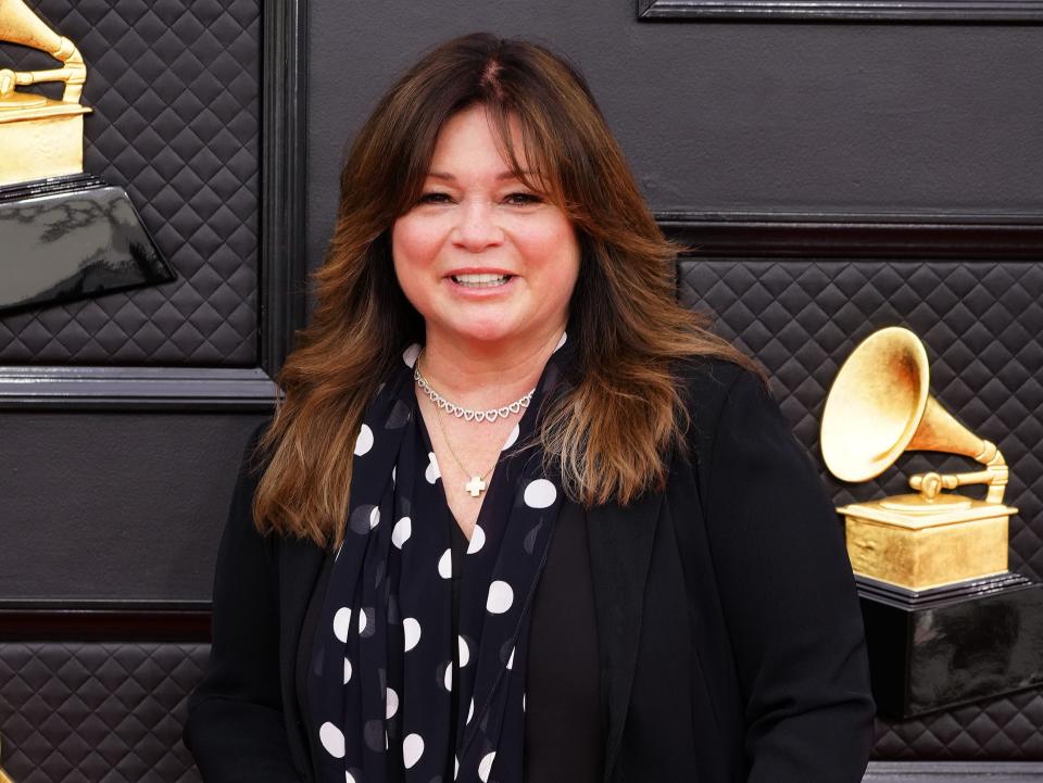 valerie bertinelli in a black and white polka dot shirt and long black cardigan blazer on the grammys red carpet, smiling and holding a clutch that reads "love"