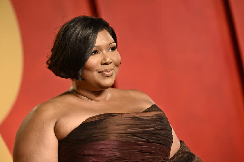 Lizzo with her hair in an updo, wearing a brown strapless gown and gloves posing against a bright red backdrop