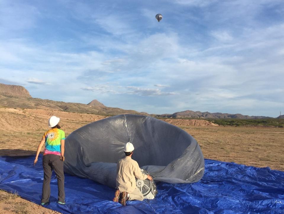 two people in white hardhats squat and stand on a blue tarp with a large grey deflated balloon watching a flying grey balloon in the distant sky