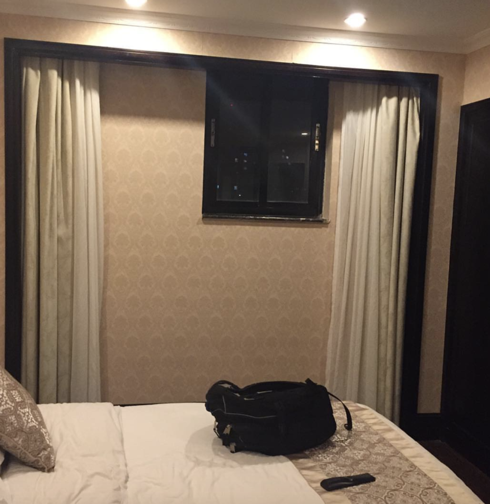 A room with a bed, a remote control on the bed, a backpack, and curtains drawn over a wall with no window