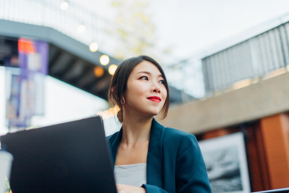 Woman with laptop looking up thoughtfully outdoors, in professional attire