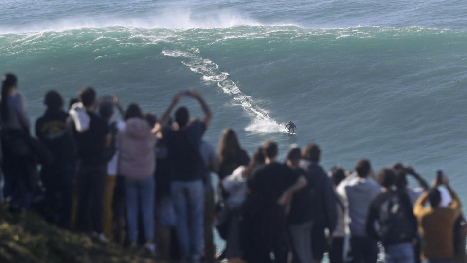 People on the top of a cliff watch surfer ride a wave during a tow surfing session at Praia do Norte or North Beach in Nazare, Portugal, Thursday, Oct. 29, 2020. A big swell generated earlier in the week by Hurricane Epsilon in the North Atlantic, reached the Portuguese west coast drawing big wave surfers to Nazare. (AP Photo/Pedro Rocha)