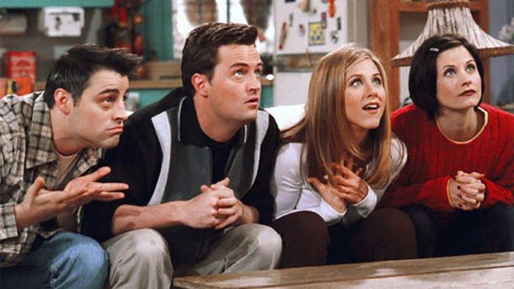 Joey, Chandler, Rachel, and Monica leaning forward looking at something intently in a scene from Friends.