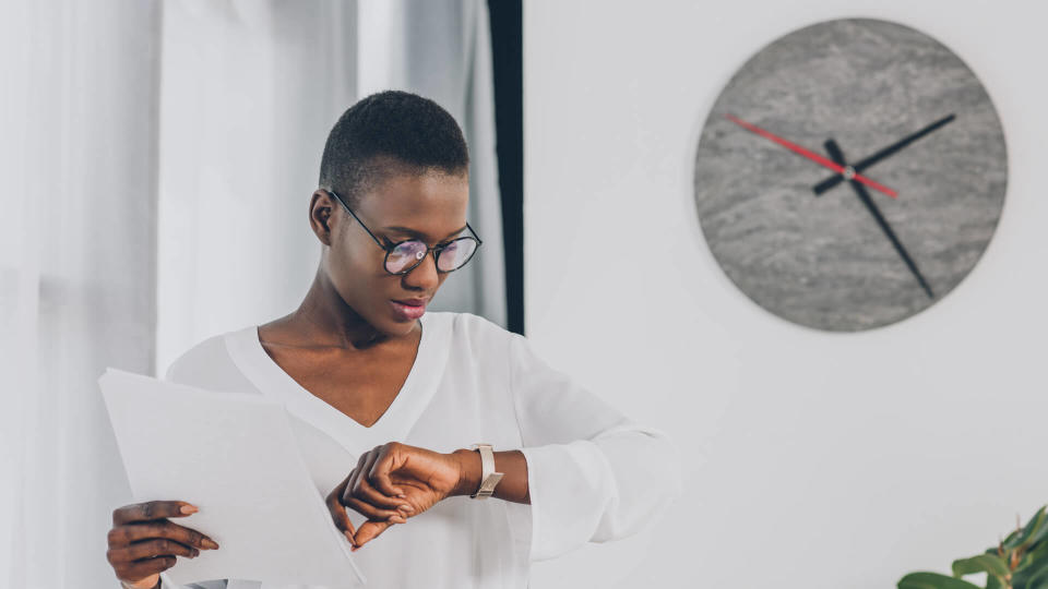 stylish african american businesswoman holding documents and checking time in office, clock on white wall.