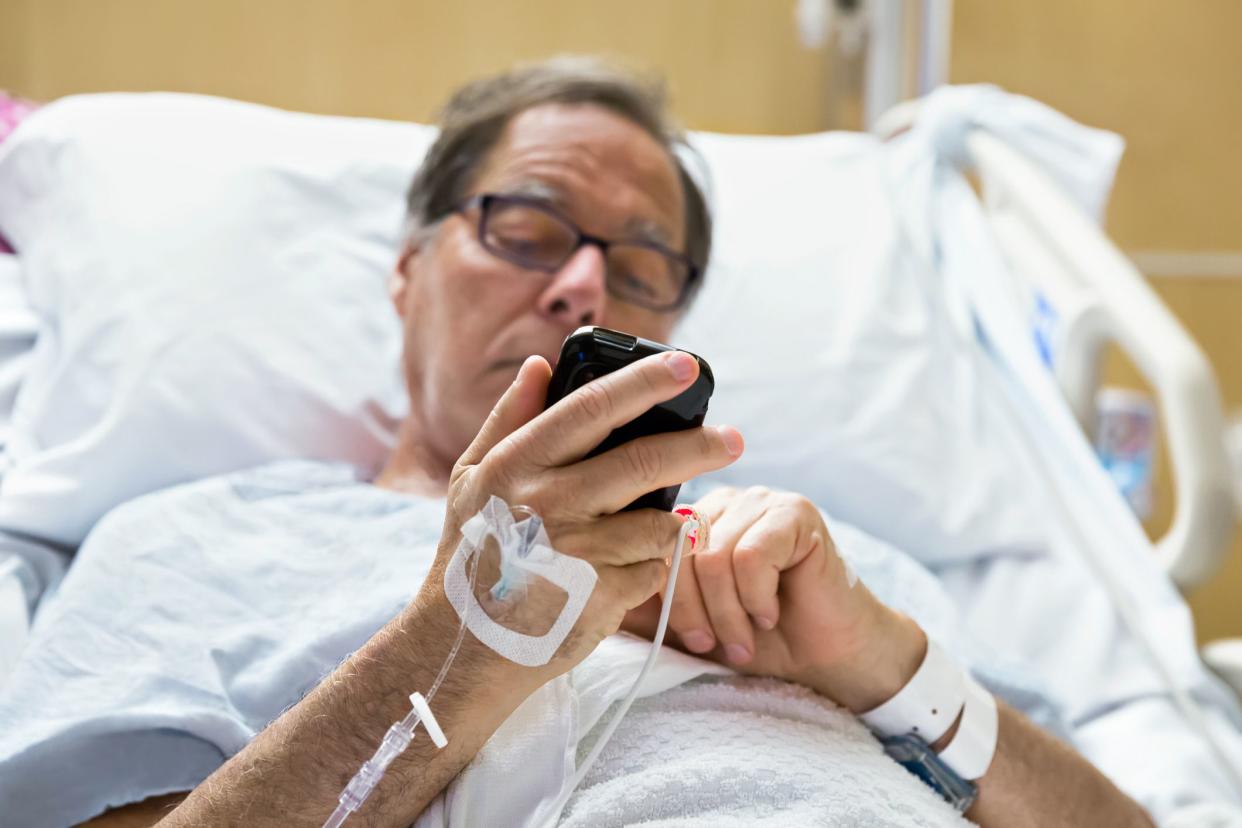 A senior male hospital patient looks at his cell phone while lying in bed.
