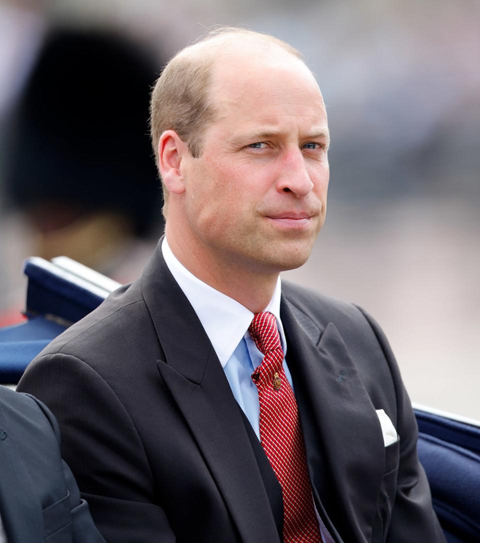 Prince William in a black suit with a red tie sitting in a carriage.