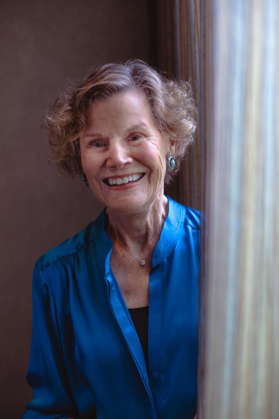 An older blonde woman in a blue shirt peeks out from behind a curtain.