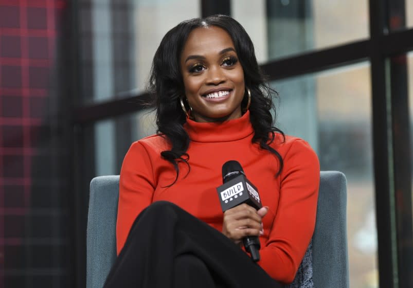 Rachel Lindsay in an orange turtleneck holding a microphone