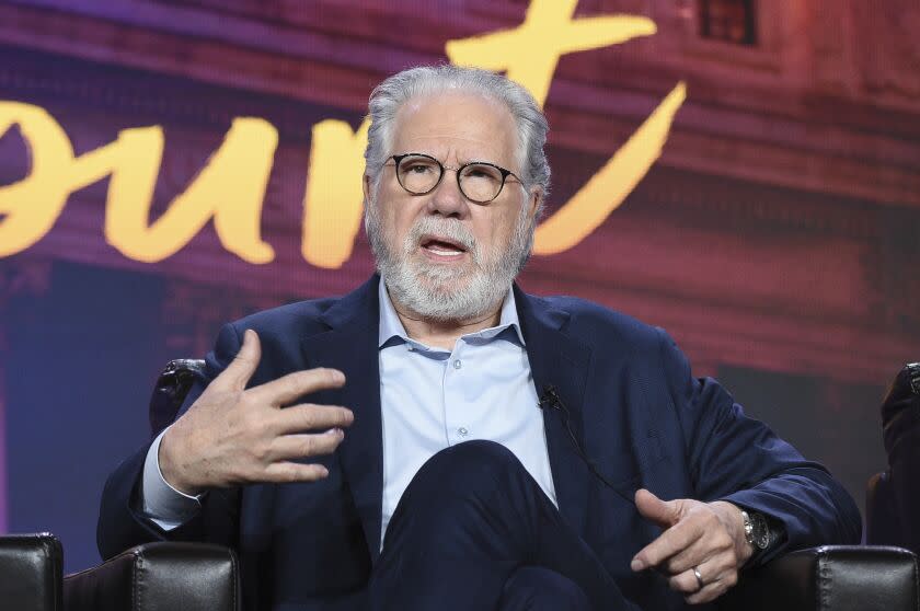 A man with white hair, a beard and glasses wearing a suit, sitting in an armchair and speaking while gesturing with his hands