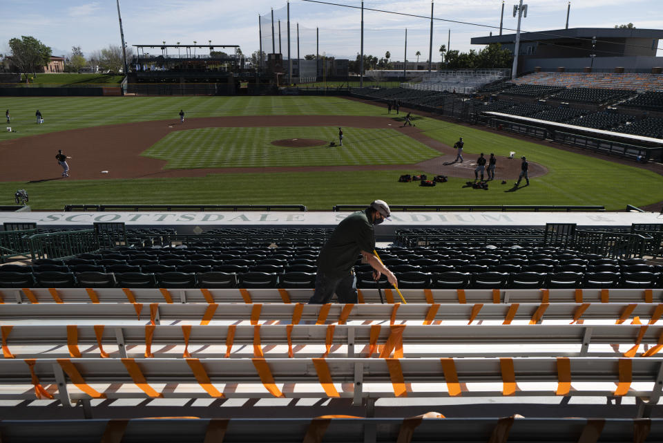 A worker cleans the stadium seats as San Francisco Giants players train during the team's spring training baseball workout in Scottsdale, Ariz., Friday, Feb. 26, 2021. The seats are taped off for social distancing for fans ahead of the team's baseball game with the Los Angeles Angels on Sunday. (AP Photo/Jae C. Hong)