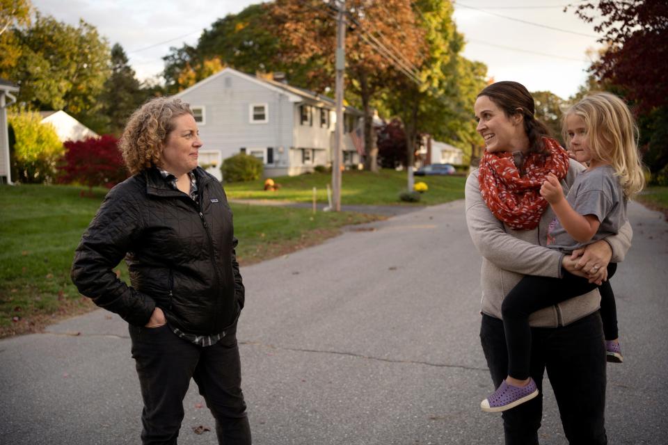 Two women stand in the residential road, chatting, and one holds a small child.