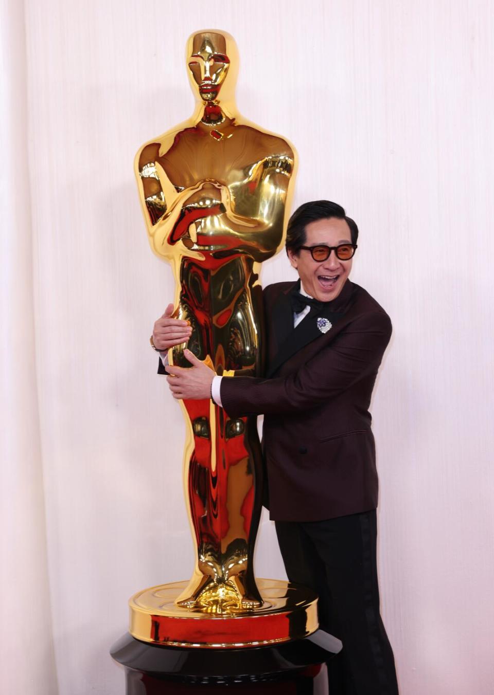 Ke Huy Quan, in a burgundy suit, hugs an Oscar statue.