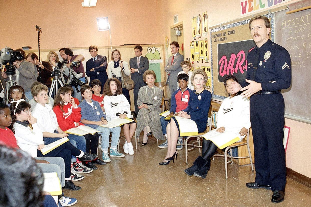 A very 1980s picture of Nancy Reagan sitting next to a uniformed and mustachioed police officer who is standing and giving a presentation in a brown-floored classroom. 