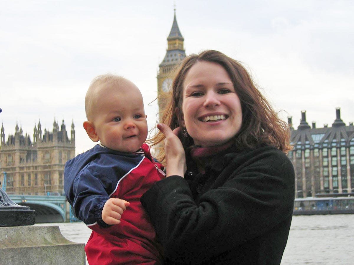 The author, with long brown hair and a black jacket, holds a smiling baby in a red and blue jacket, in front of Big Ben in London