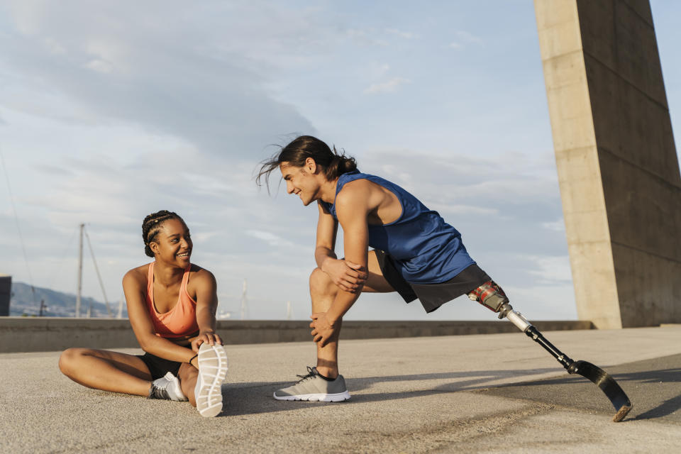 A woman and a man with a prosthetic leg are stretching outdoors, smiling at each other. They are dressed in casual athletic wear