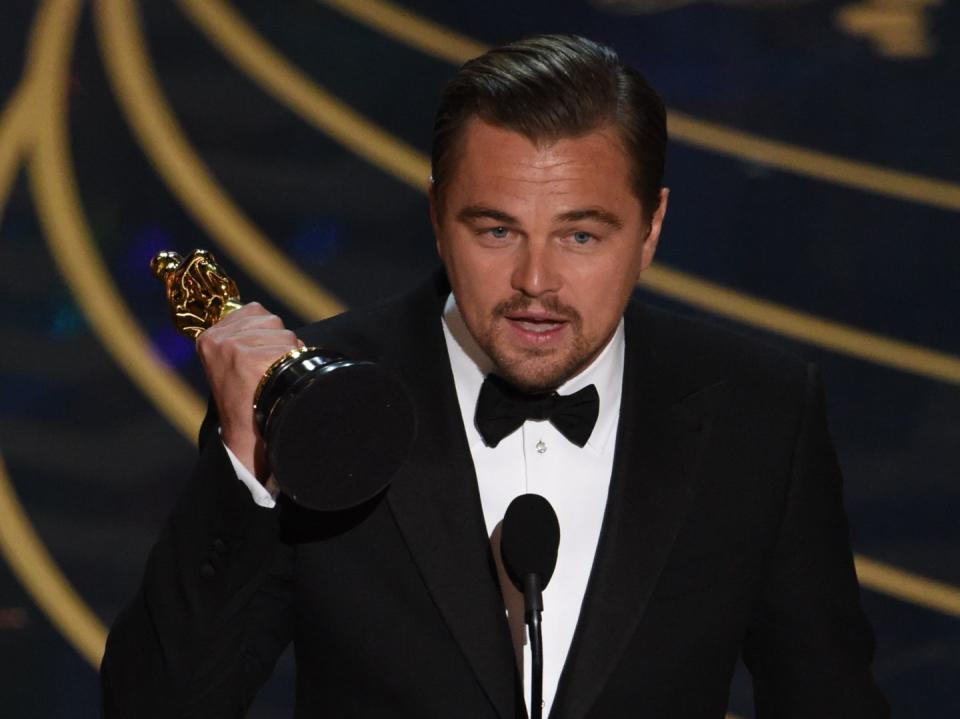 leonardo dicaprio wearing a black tuxedo, standing on a stage and holding an oscar statuette while speaking into a microphone