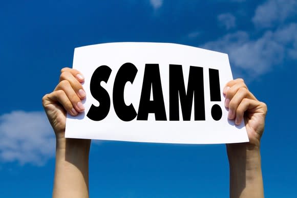 Two hands are holding up a sign on which is printed the word "scam" with an exclamation mark.