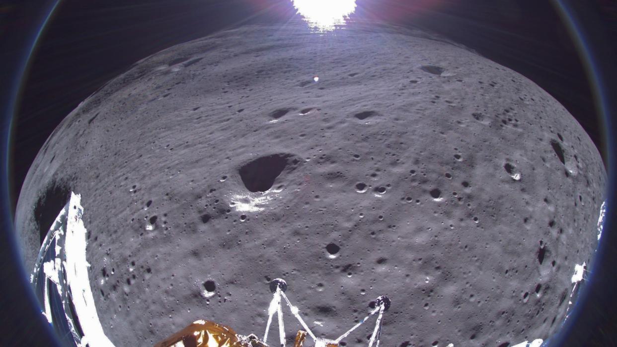  A fisheye photo showing parts of a gold and silver lunar lander in the foreground, with the moon's gray dirt and the bright sun in the background. 