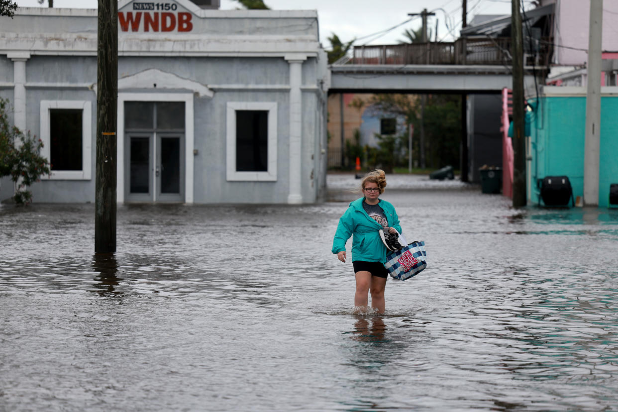 Brooke Voegtle, in shorts and bare legs, with a puffer jacket and a shopping bag, walks through shin-deep floodwater in front of a building marked WNDB.