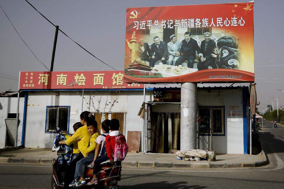 In this Sept. 20, 2018, photo, an Uighur woman uses an electric-powered scooter to fetch school children as they ride past a picture showing China's President Xi Jinping joining hands with a group of Uighur elders at the Unity New Village in Hotan, in western China's Xinjiang region. While thousands of Uighur Muslims across China’s Xinjiang region are forced into re-education camps, China’s fledgling vision for ethnic unity is taking shape in a village where Han Chinese work and live alongside Uighur minorities. (AP Photo/Andy Wong)