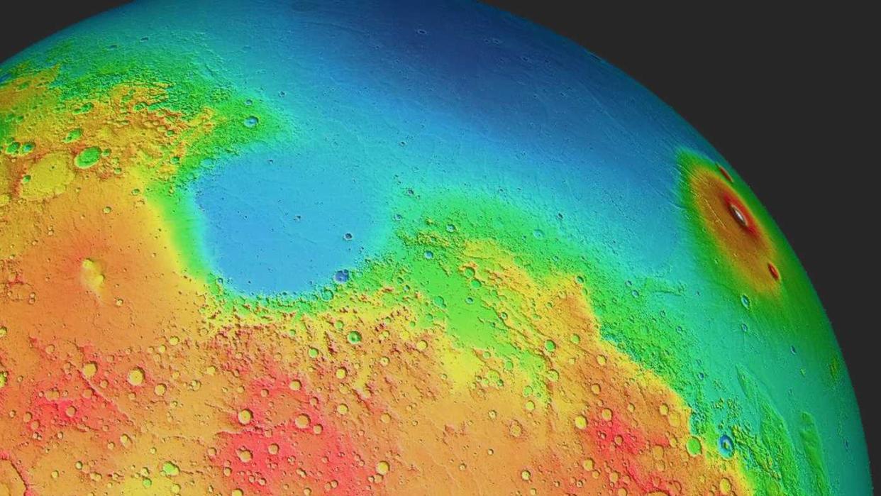  a topographical map of mars from a position seen in space. a huge basin in blue is at the top with higher altitude regions surrounding it, in green, red and orange. 