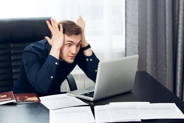 A young businessman at his desk, clutching his head and staring wide-eyed at his computer screen.