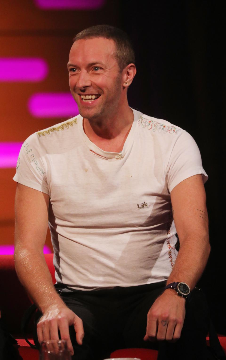 Chris Martin sitting, smiling in a white t-shirt with decorative collar, wearing a wristwatch on a talk show