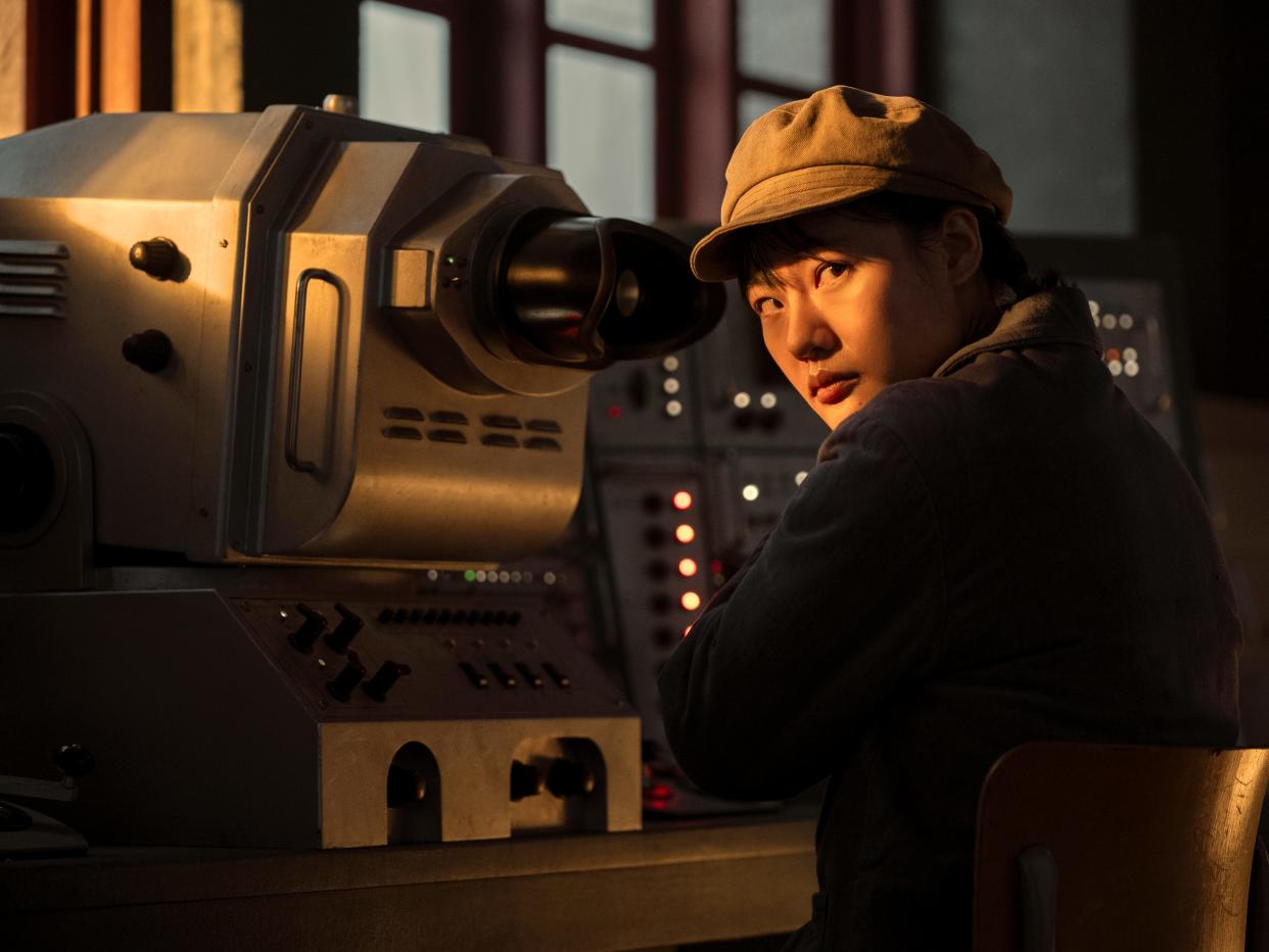 zine tseng as ye wenjie in three body problem. she's a young woman sitting in front of a control panel, wearing a brown hat with a brim, and a blue jacket. bathed in warm light, she looks over her shoulder