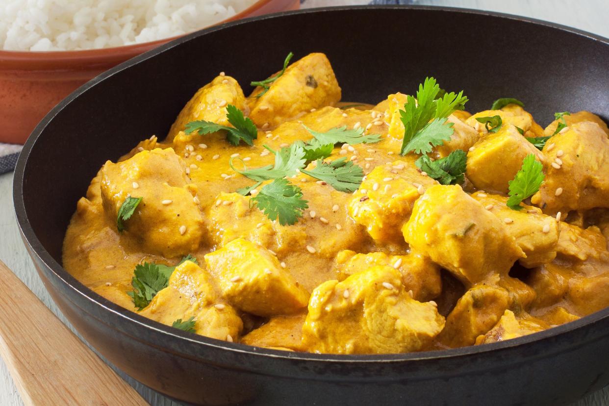 Closeup of chicken curry in a black ceramic bowl, next to a terracotta bowl of rice and a wooden spoon on a table