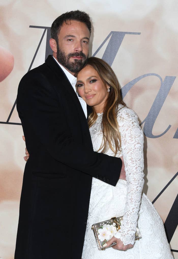 Ben Affleck and Jennifer Lopez embrace on a red carpet. Affleck wears a black coat, and Lopez wears a long-sleeve, lace gown while holding a floral clutch