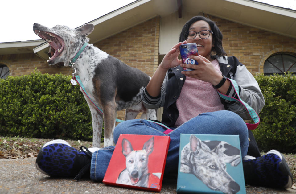 In this photo made Monday, March 2, 2020, Danielle Moore looks at her phone as she poses for photos with Kana and paintings of the pet Australian cattle dog in Dallas. In the dog-eat-dog world of online shopping, Chewy has an unusual plan to fend off Amazon: turning pets into works of art. The online pet shop surprises customers with oil paintings of their furry friends, a move the company says wins them customers for life. (AP Photo/LM Otero)