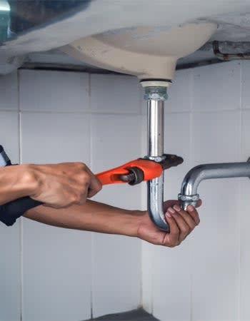 A pair of hands use a tool to fix plumbing under the sink.