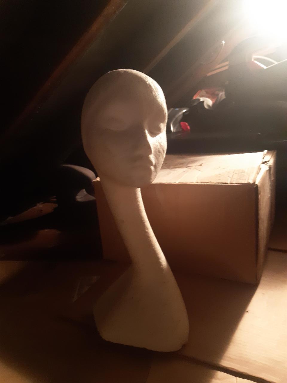 A mannequin head used for hats or wigs is stored in a dimly lit, cluttered space