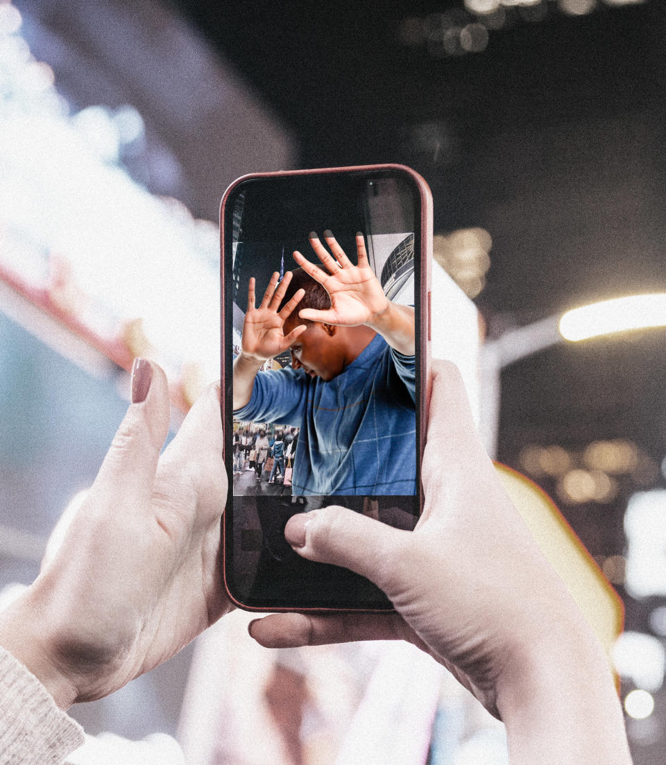 A phone screen's camera shows a man shielding his face as someone tries to take a picture of him