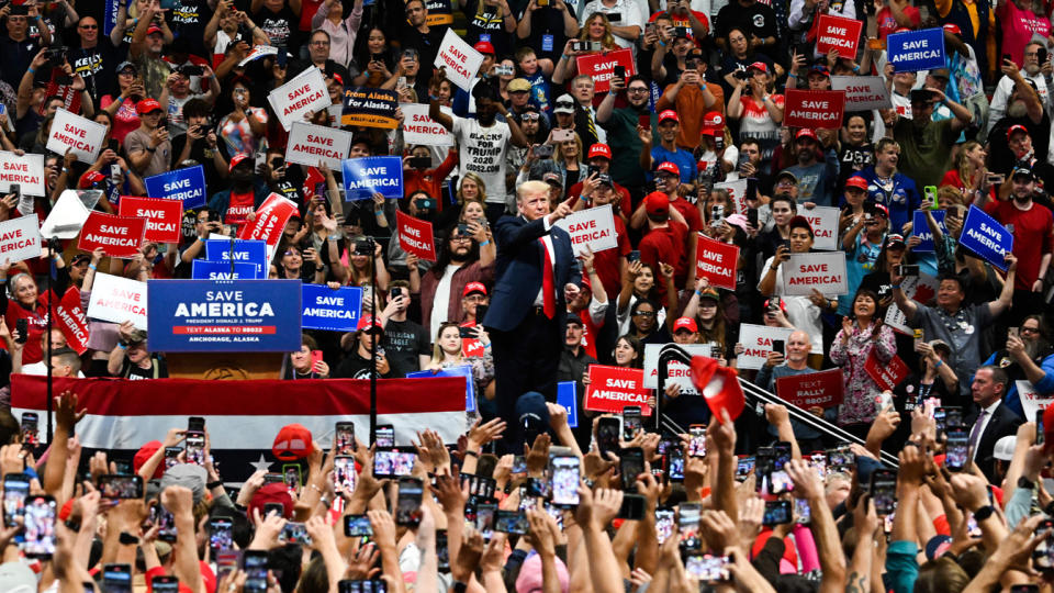 Donald Trump stands onstage pointing amid throngs of supporters who carry signs that read Save America.