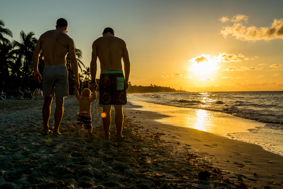 Two adults and a child holding hands, walking on a beach during sunset. Sunlight reflects on the water and sand