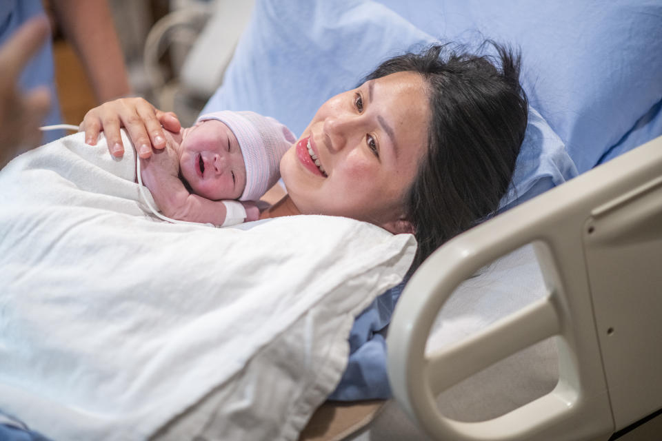 A young Asian mother lays in her hospital bed in the delivery room after giving birth to her son.  She has him laying on her chest as the share an intimate moment together and some skin-to-skin time.  She is wearing a blue hospital gown and has a white blanket draped over the newborn.