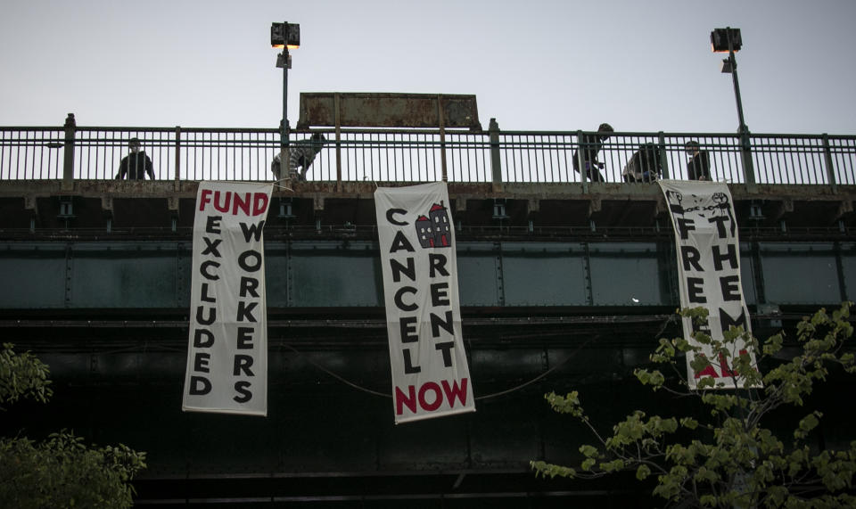 People unfold banners from a subway platform during a vigil memorializing more than sixty persons who died from COVID-19 and were associated with Make the Road New York (MRNY), a support organization for immigrant and working class communities, Thursday May 21, 2020, in Corona Plaza, Queens, N.Y. With support from elected state and city officials, MRNY has launched a campaign called #Recovery4All, demanding Governor Cuomo and Mayor de Blasio create "a $3.5 billion Excluded Workers' Fund, cancel rent, and release at risk people from prisons and detention centers" to address the disproportionate effects from the pandemic in Queens. (AP Photo/Bebeto Matthews)