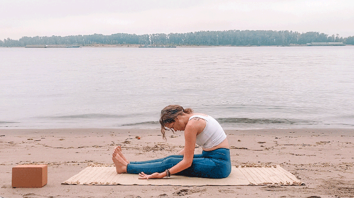 Woman sitting on a yoga mat at the beach leaning her chest toward her thighs in a Yin Yoga pose