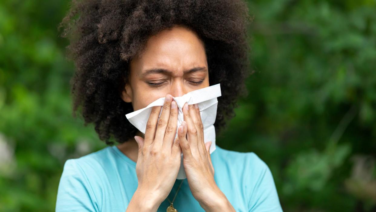  young black woman blowing her nose into a tissue while standing outside near some trees 