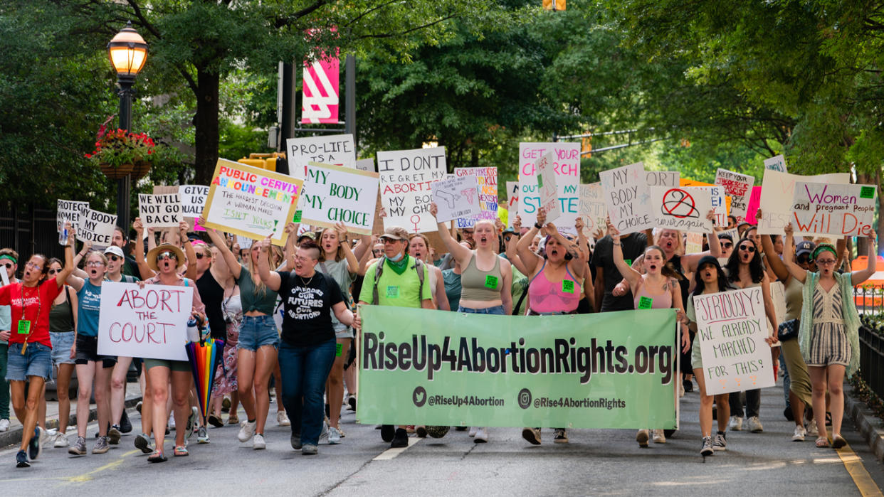 Protesters, mainly women, march in the street carrying banners saying such things as: Abort the Court; My Body, My Choice; Women and pregnant people Will Die; and Get Your Church Out of My State.