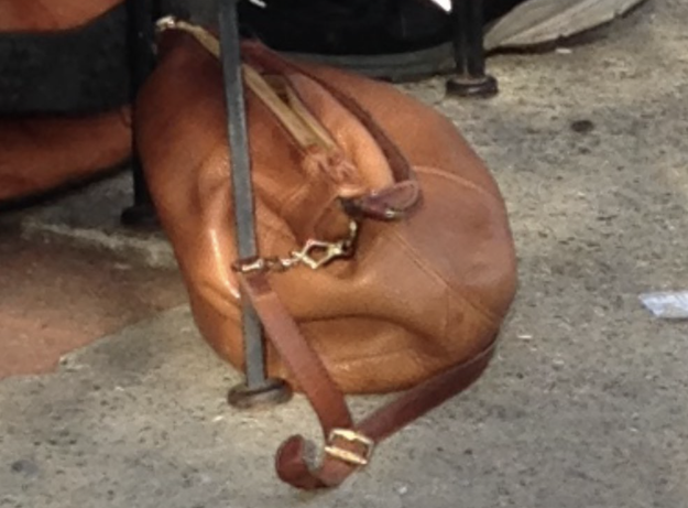 A worn brown leather bag with a long strap resting on pavement