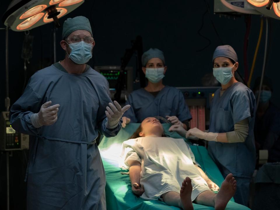 ellie in the last of us laying, unconscious, on a surgical table, wearing only a hospital gown and with bare feet. around her stands a team of doctors and nurses, all wearing blue srubs, caps, face masks, and gloves. the doctor in the front is explaining himself to someone, holding his hands up