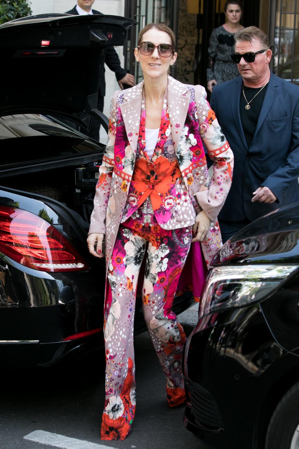 Celine in an all-over floral print suit. The matching blazer, blouse, and pants had pink red and orange flowers. She is wearing brown sunglasses.